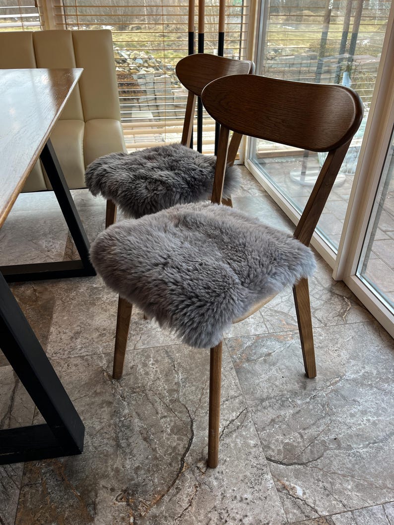 May include: Two wooden chairs with gray sheepskin seat covers. The chairs have curved backs and slender legs. The sheepskin covers add a touch of texture and comfort to the seating. The chairs are placed near a window.