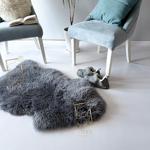 May include: A grey sheepskin rug is on a white surface, with a pair of grey slippers nearby. Two light blue chairs with white legs are in the background, one with a fluffy white cushion and a book, the other with a blue cushion.