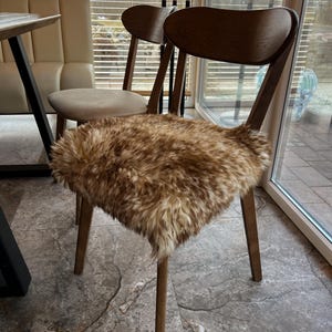May include: A wooden chair with a curved back and four legs. The seat is covered with a brown and tan faux fur cushion. Another chair with a beige seat is visible in the background. The chairs are in a room with a window.