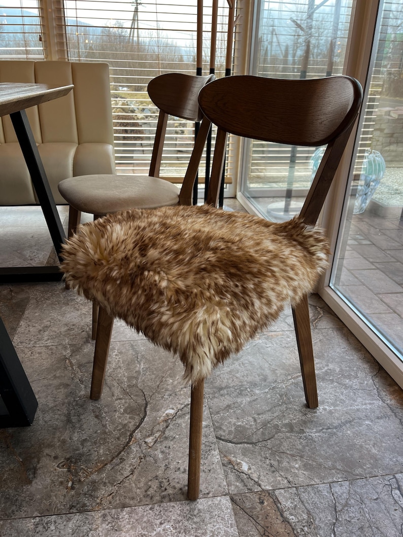 May include: A wooden chair with a curved back and four legs. The seat is covered with a brown and tan faux fur cushion. Another chair with a beige seat is visible in the background. The chairs are in a room with a window.