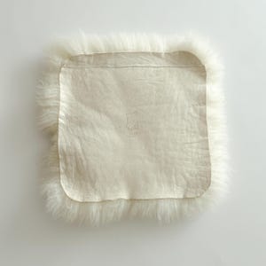 May include: A square, off-white sheepskin rug with a soft, fluffy border. The center features a smooth, light-colored leather backing. The rug's design is simple and natural, with a focus on texture and neutral tones. The overall aesthetic is cozy and inviting.