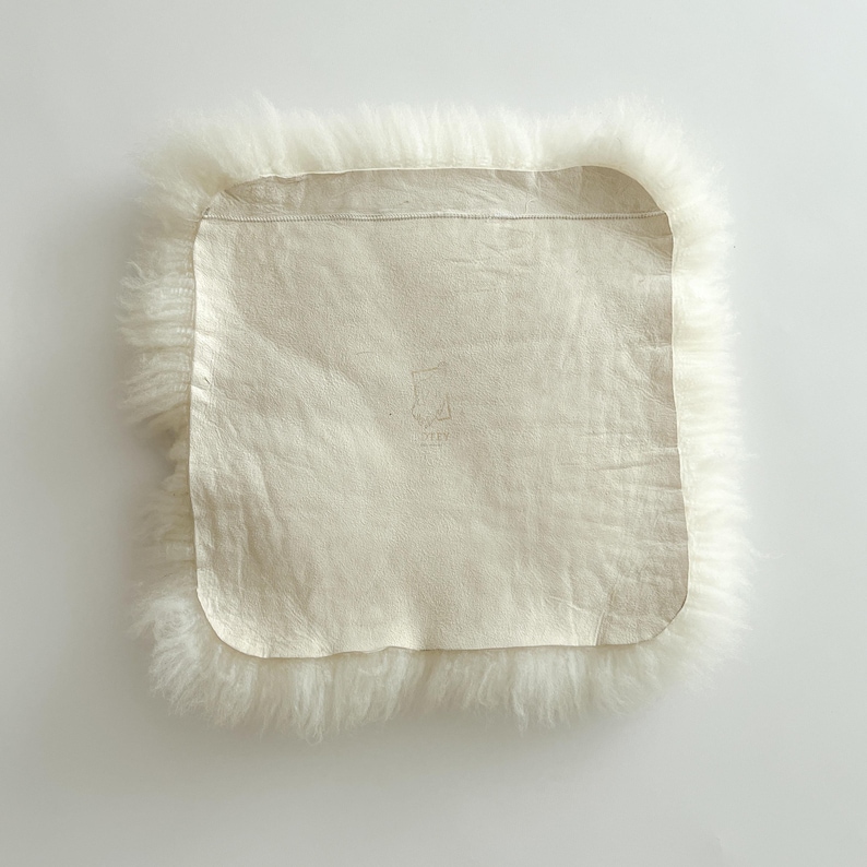 May include: A square, off-white sheepskin rug with a soft, fluffy border. The center features a smooth, light-colored leather backing. The rug's design is simple and natural, with a focus on texture and neutral tones. The overall aesthetic is cozy and inviting.