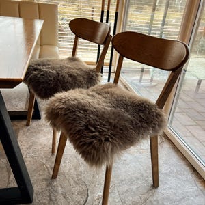May include: Two wooden chairs with curved backs and light brown fur seat covers. The chairs have four slender legs and are positioned near a window with blinds. The chairs are in a well-lit room with a table visible on the left.