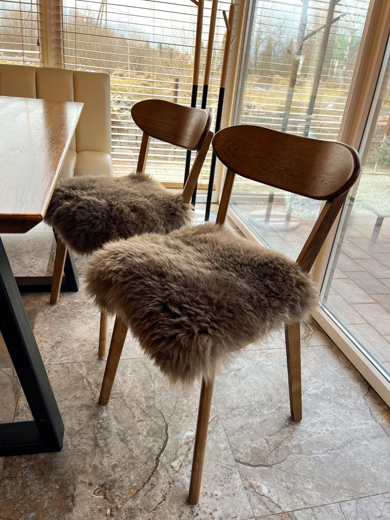 May include: Two wooden chairs with curved backs and light brown fur seat covers. The chairs have four slender legs and are positioned near a window with blinds. The chairs are in a well-lit room with a table visible on the left.