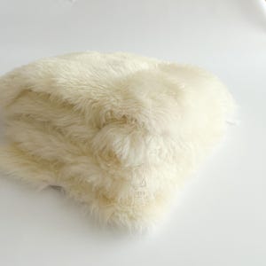 May include: Cream-colored, fluffy sheepskin rug. The rug has long, soft fibers and a dense texture. The sheepskin is folded over itself, creating a layered appearance. The background is a plain white surface.