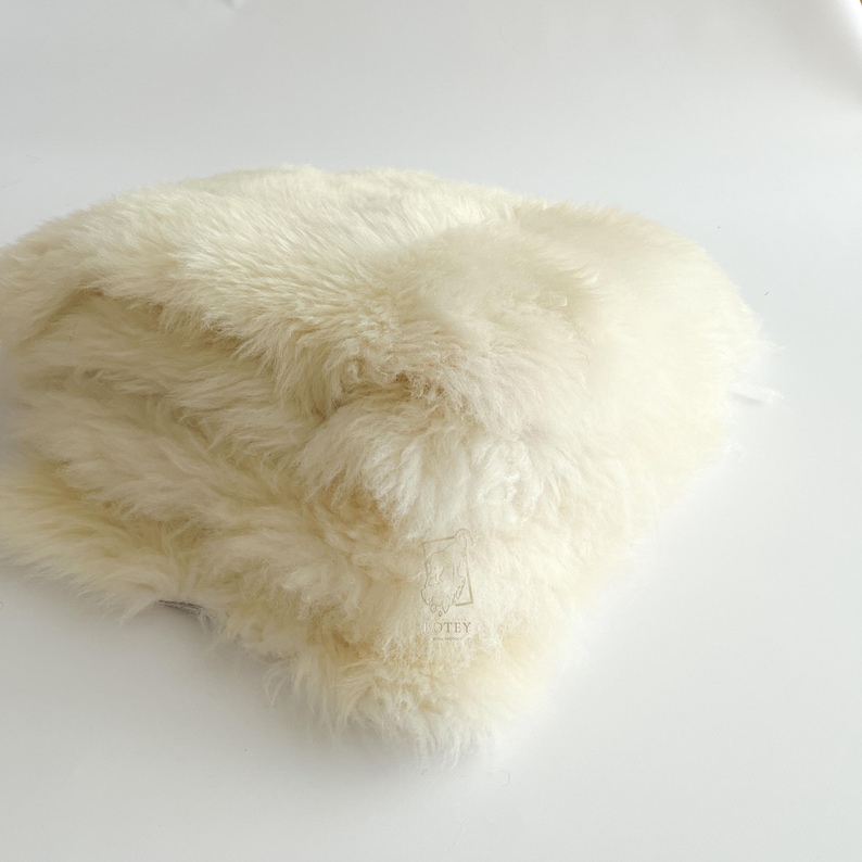 May include: Cream-colored, fluffy sheepskin rug. The rug has long, soft fibers and a dense texture. The sheepskin is folded over itself, creating a layered appearance. The background is a plain white surface.