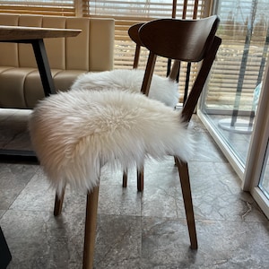 May include: Two wooden chairs with light brown legs and dark brown backs. Each chair has a fluffy, white sheepskin seat cover. The chairs are in a well-lit room with a table and a window with blinds.