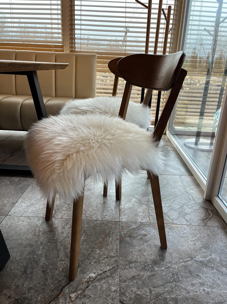 May include: Two wooden chairs with light brown legs and dark brown backs. Each chair has a fluffy, white sheepskin seat cover. The chairs are in a well-lit room with a table and a window with blinds.