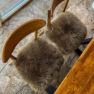 May include: Two wooden chairs with curved backs and light brown sheepskin seat cushions. The chairs are positioned near a window and a wooden table, creating a warm and inviting seating area. The sheepskin cushions add a touch of comfort and texture.