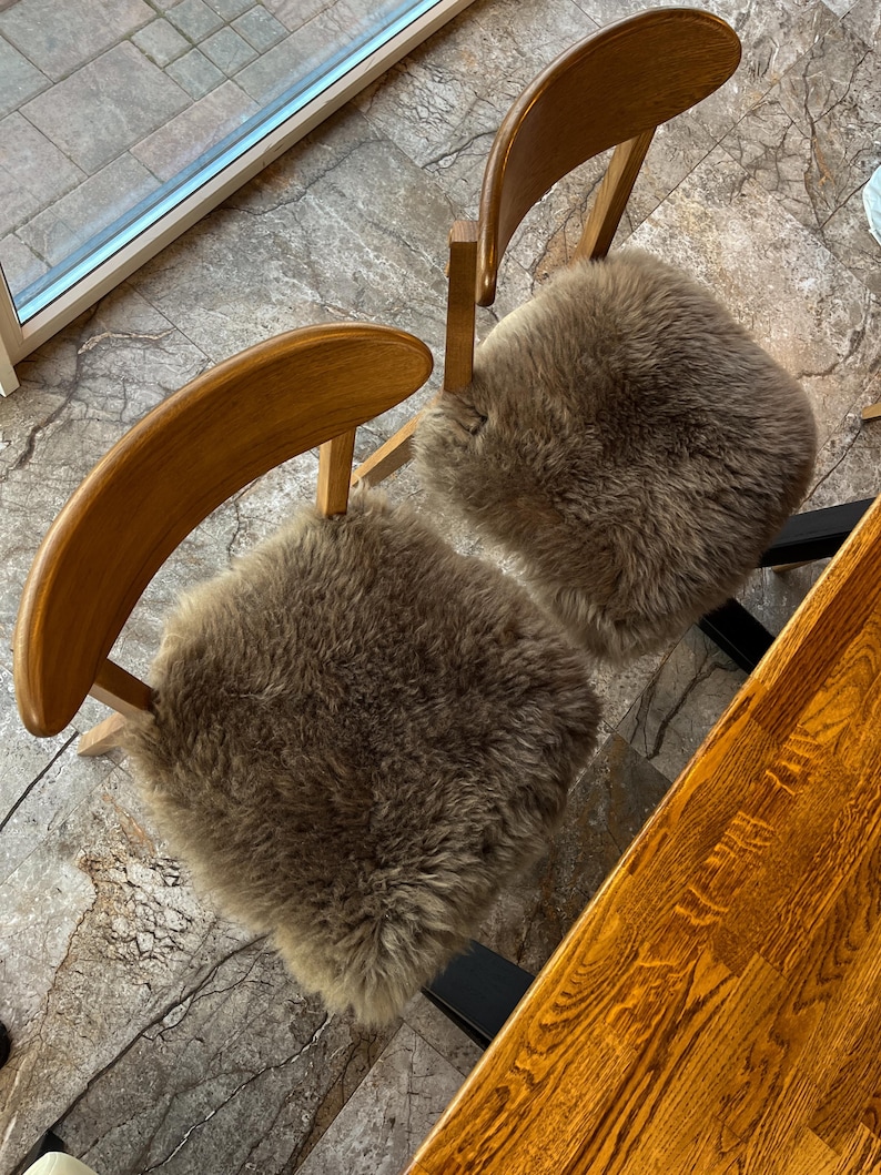May include: Two wooden chairs with curved backs and light brown sheepskin seat cushions. The chairs are positioned near a window and a wooden table, creating a warm and inviting seating area. The sheepskin cushions add a touch of comfort and texture.