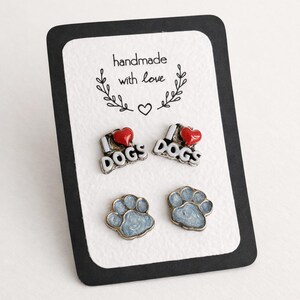 Può includere: Due paia di orecchini a lobo in metallo su un cartoncino bianco con bordi neri. Un paio presenta la scritta I heart DOGS con un cuore rosso. Il secondo paio raffigura impronte di zampe azzurre. Il testo sul cartoncino è handmade with love.