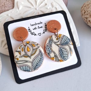 Può includere: Orecchini fatti a mano con un design floreale. Gli orecchini hanno una parte superiore in legno e un ciondolo circolare con sfondo crema e motivi floreali blu, gialli e neri. Il testo "handmade with love" è stampato sulla carta.