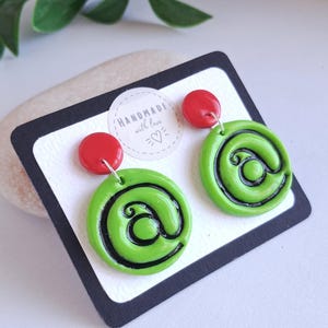 Puede incluir: Un par de pendientes colgantes con un poste circular rojo brillante conectado a un colgante circular verde lima más grande. Los colgantes verdes tienen un diseño de espiral negro que se asemeja al símbolo de arroba, sobre una tarjeta blanca.