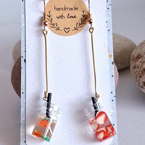 May include: Handmade earrings featuring miniature clear bottles filled with colorful fruit and straws. One earring has green and orange fruit, the other has red fruit. The earrings hang from gold-colored hooks and long bars. The backing card says "handmade with love".