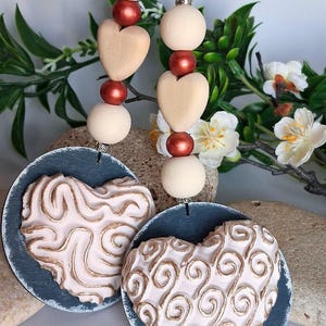 May include: Decorative hanging ornaments featuring heart and swirl designs. Each ornament has a blue-gray circular base with a raised, textured heart design in cream and gold. Cream-colored beads and heart-shaped accents are strung above.