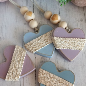 May include: Four wooden hearts with lace trim, painted in shades of blue and purple. The hearts are strung on a cord with wooden beads.