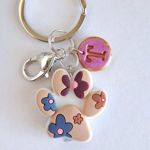 Può includere: Un portachiavi argentato con un ciondolo a forma di zampa con un design floreale nei toni del beige, blu e marrone. Include un ciondolo rotondo con la lettera "T" in un cerchio rosa. Il portachiavi ha un moschettone.