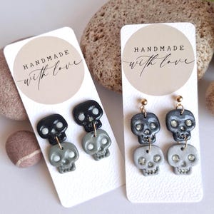 May include: Two pairs of skull-shaped drop earrings displayed on white cards with the text HANDMADE with love. One pair features solid black and grey skulls, while the other pair features metallic charcoal and silver skulls with glittery accents.