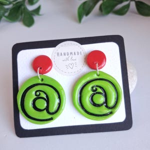Op de afbeelding: Een paar hangende oorbellen met een felrode ronde knop bevestigd aan een grotere limoengroene ronde hanger. De groene hangers zijn voorzien van een zwart apenstaartje. De oorbellen worden gepresenteerd op een kaart met de tekst HANDMADE with love.