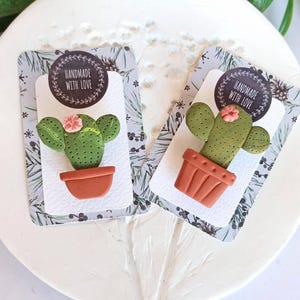 Può includere: Due ornamenti a forma di cactus fatti a mano con fiori rosa in vasi di terracotta. Ogni ornamento è su una carta decorativa con un motivo invernale e un adesivo "Handmade with Love".