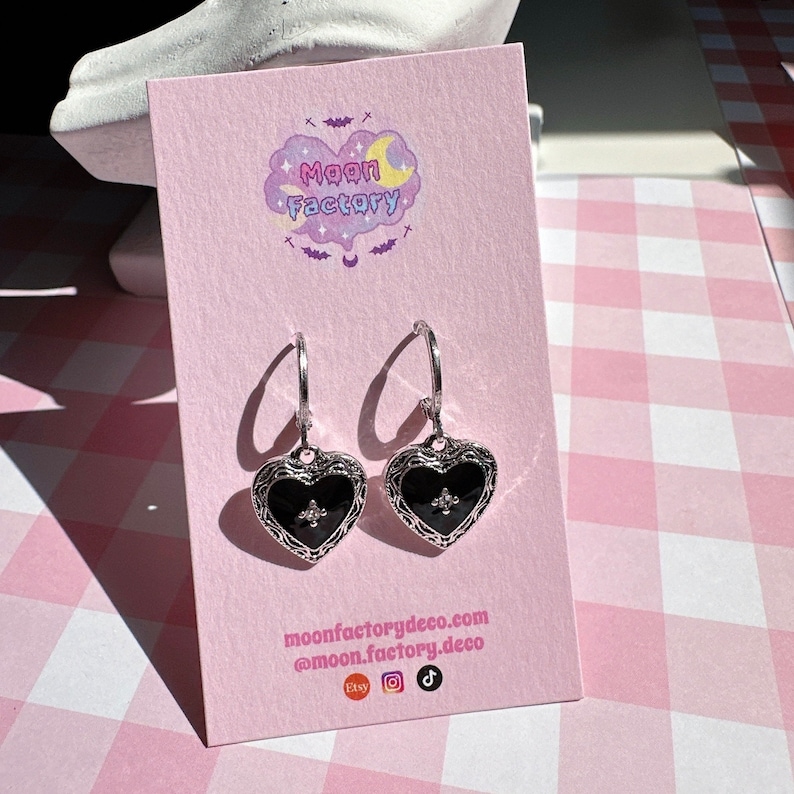 May include: A pair of silver hoop earrings with black heart charms. Each charm has a silver fleur-de-lis design in the center. The earrings are displayed on a pink card with the text "Moon Factory" and the website address "moonfactorydeco.com".