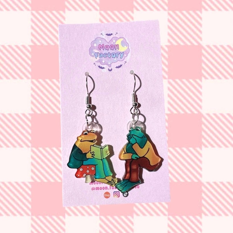 May include: A pair of dangle earrings featuring a green and brown frog reading a book and a green frog sitting with its arms crossed. The earrings are silver and have a clear background. The earrings are on a purple card with the text "Moon Factory" and social media handles.