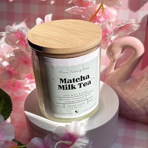 May include: A clear glass candle jar with a wooden lid, labeled "Matcha Milk Tea." The label lists soy wax blend, matcha, steamed milk, and orange blossom. The candle is 10 oz and hand-poured in SoCal. The candle sits on a white pedestal, surrounded by pink flowers.