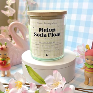 May include: A clear glass candle jar with a light green wax fill and a bamboo lid. The label features a light green gingham pattern, the text Melon Soda Float, Moon Factory Deco, Soy Wax Blend, Honeydew Melon, Fizzy Bubbles, Cream Soda, 10oz, 283g.