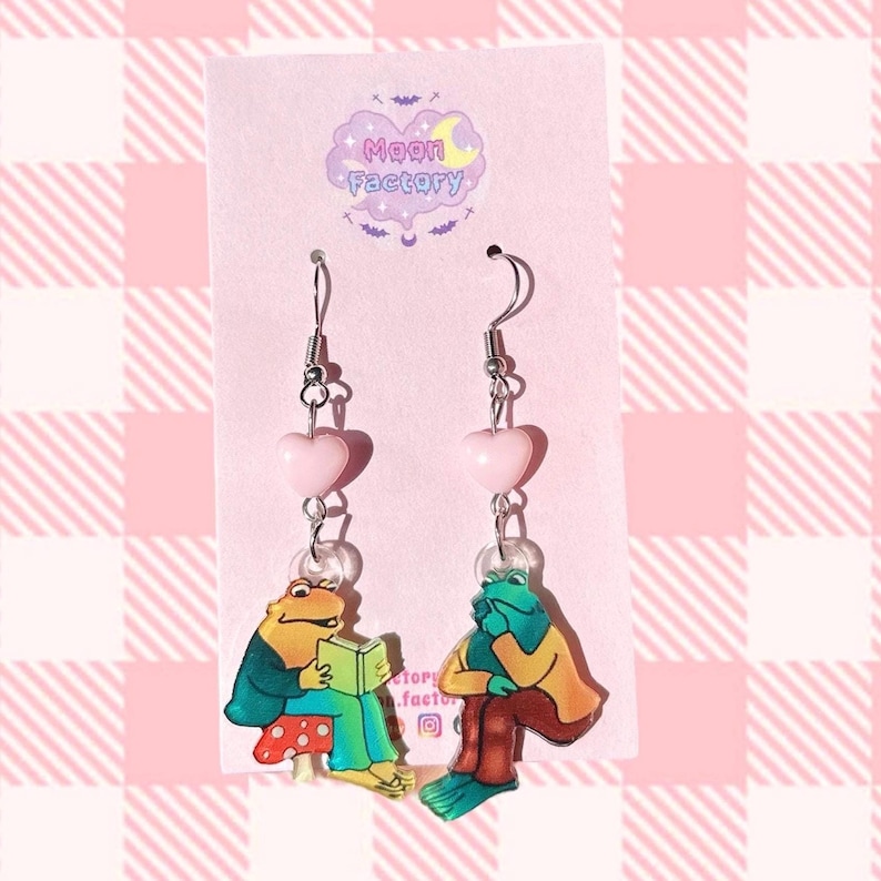 May include: A pair of dangle earrings featuring a pink heart charm and a green frog charm. The frog charm is sitting on a mushroom and holding a book. The earrings are labeled "Moon Factory".