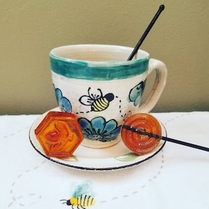 May include: A white teacup with a green rim and a bee design, a saucer, and two honey candies. One candy is a hexagonal shape and the other is a round lollipop.