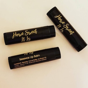 May include: Three black lip balm tubes with gold lettering. The tubes are labeled "Howe Sweet It Is" and "Beeswax Lip Balm". The ingredients are listed on the bottom tube.