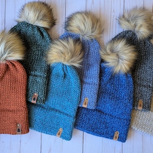 May include: A collection of knitted winter hats in various colors, including rust, teal, and blue, each topped with a fluffy faux fur pom-pom. The hats have a leather tag detail and are arranged on a light wood surface.