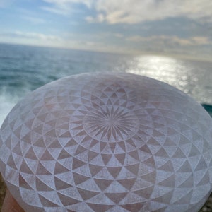 May include: A pale, round object with an intricate geometric pattern carved into its surface. The design features a central spiral surrounded by triangular shapes. The background shows the sea and sky.