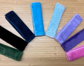 May include: A collection of eight rectangular, plush fabric wristbands arranged in a fan shape. The colors include dark green, black, navy blue, bright blue, light blue, lavender, deep purple, and bright pink, all featuring a soft, fuzzy texture.