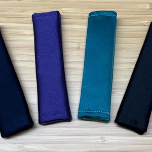 May include: Four fabric wrist straps in different colors: black, purple, teal, and gray. The straps are all the same size and shape, and they have a smooth, soft texture.