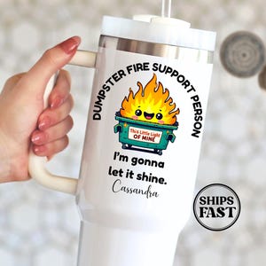 May include: White tumbler with a handle, featuring the text "DUMPSTER FIRE SUPPORT PERSON" and a cartoon dumpster fire graphic. The tumbler also includes the text "I'm gonna let it shine" and a name.