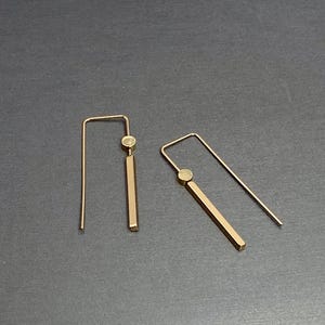May include: A pair of gold-colored earrings. Each earring features a straight, rectangular bar suspended from a small, round accent piece and a long, thin hook. The earrings are displayed on a dark gray surface.