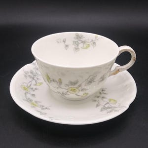 May include: A white teacup and saucer with a floral pattern in shades of gray and yellow. The cup and saucer have a textured surface.