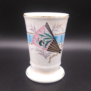 May include: A white ceramic cup with a blue and gold band around the top. The cup has a hand-painted design of a pink and black fan with green leaves and a gold stem. The cup has a gold rim and a white base.