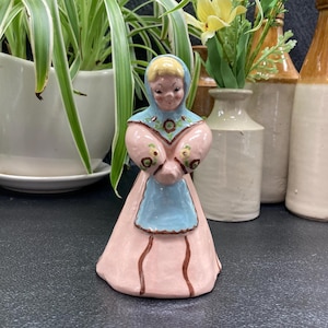 May include: A ceramic figurine of a woman wearing a pink dress, blue apron, and a white head covering. She has brown hair and brown eyes. The figurine is decorated with brown and yellow floral details.