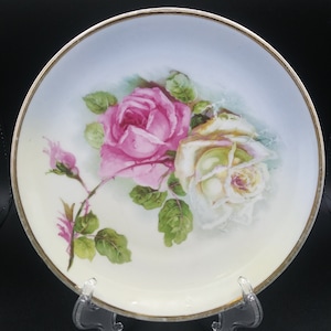 May include: A white porcelain plate with a gold rim features a hand-painted design of two roses. One rose is pink and the other is white.