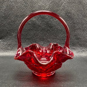 May include: A vibrant red glass basket with a handle. The basket features a ruffled edge and a textured surface. The handle curves over the basket. It rests on a small, circular base. The glass is transparent.
