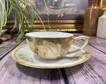May include: A vintage teacup and saucer set. The cup and saucer are white with a gold rim and floral rose design. The teacup has a handle and sits on a matching saucer. The set is displayed on a wooden surface.