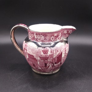 May include: A silver-plated creamer with a pink and white floral design and a scene of ships. The creamer has a handle and a spout.