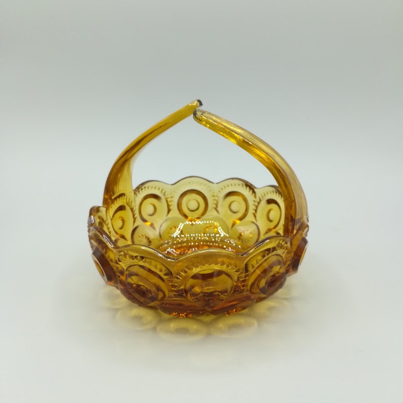 May include: Amber-colored glass basket with a handle. The basket features a repeating pattern of circles and crescent shapes around the rim and on the base. The handle arches gracefully from side to side. The glass has a glossy finish.