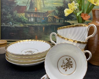 May include: A set of white porcelain teacups and saucers with gold trim. The teacups have a spiral design and a gold rose emblem inside. The saucers are stacked, and the background includes a painting of a windmill and flowers.