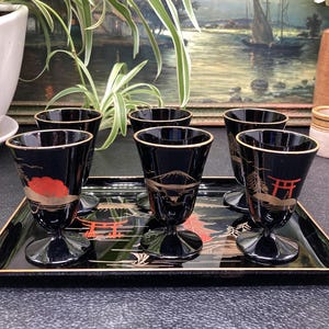 May include: Set of six black and gold sake cups with Japanese designs on a black lacquer tray. The cups feature gold accents and red details, including a red mountain and a red torii gate.