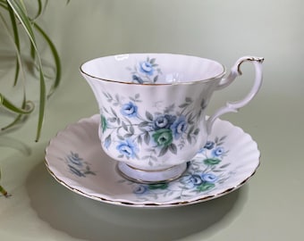 May include: A vintage white teacup and saucer set, decorated with blue and green rose motifs. The teacup and saucer have gold edging. The teacup and saucer are likely made of fine bone china.