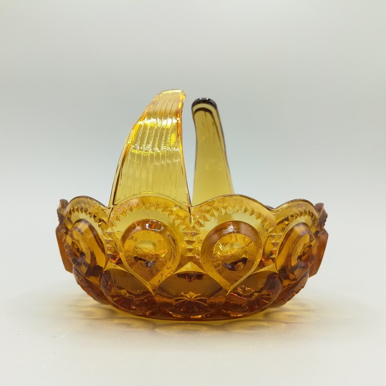 May include: Amber-colored glass basket with a handle. The basket features a star and circle pattern around the rim and on the base. The handle is arched and has vertical ridges. The glass has a glossy finish.