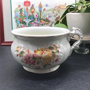 May include: A white ceramic chamber pot with a gold rim and ornate handle. The pot is decorated with a floral pattern featuring pink, yellow, and orange roses. The pot has a wide opening and a pedestal base.